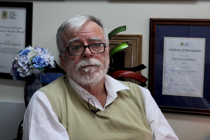 A man with white hair, moustache and beard sits in front of framed degrees, with neutral expression.