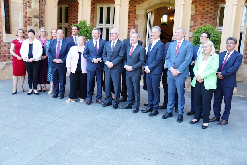 A group of 18 well dressed people standing in a row and smiling for the cameras. 