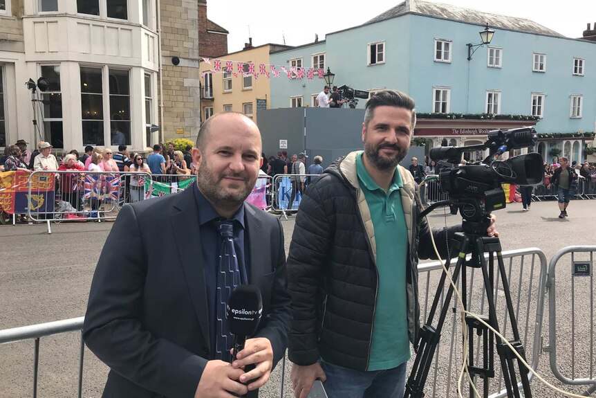 A man in a suit holds a microphone, and stands next to a camera man on a street lined with royal wedding rans behind baricades