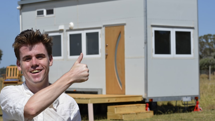A young person with short hair, wearing a long-sleeve white T-shirt, standing in front of a tiny home.