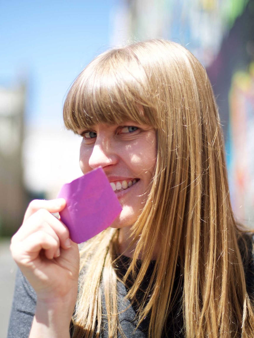 Tight portrait shot of a woman holding a purple Post-It note up to her face.