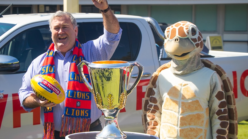 a man with his fist in the air standing next to the AFL premiership cup and a turtle mascot