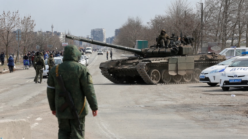 Soldiers sit atop a tank that is on a wide road near parked police cars, with a line of people looking from some distance.