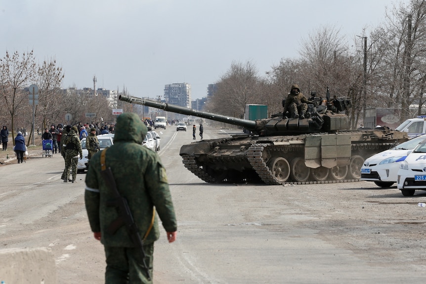 Soldiers sit atop a tank that is on a wide road near parked police cars, with a line of people looking from some distance.