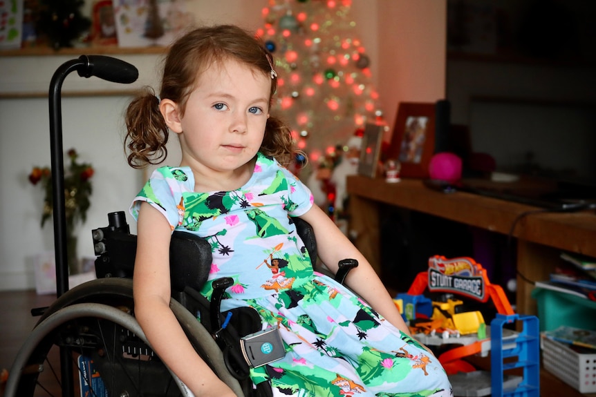 A young girl wearing a dress with green dinosaur smiles at the camera from her living room.