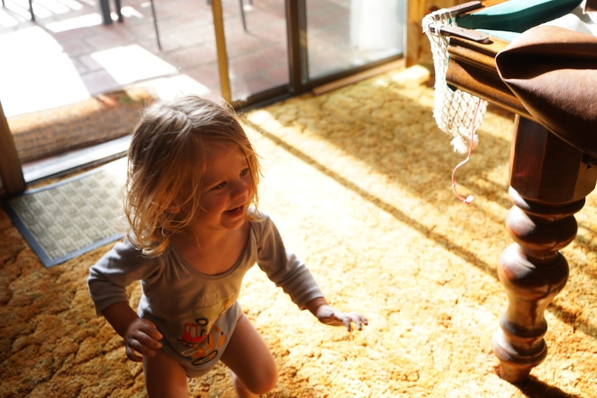 A toddler runs in a lounge room near a pool table, with an expression of glee on his face. He has blonde hair.