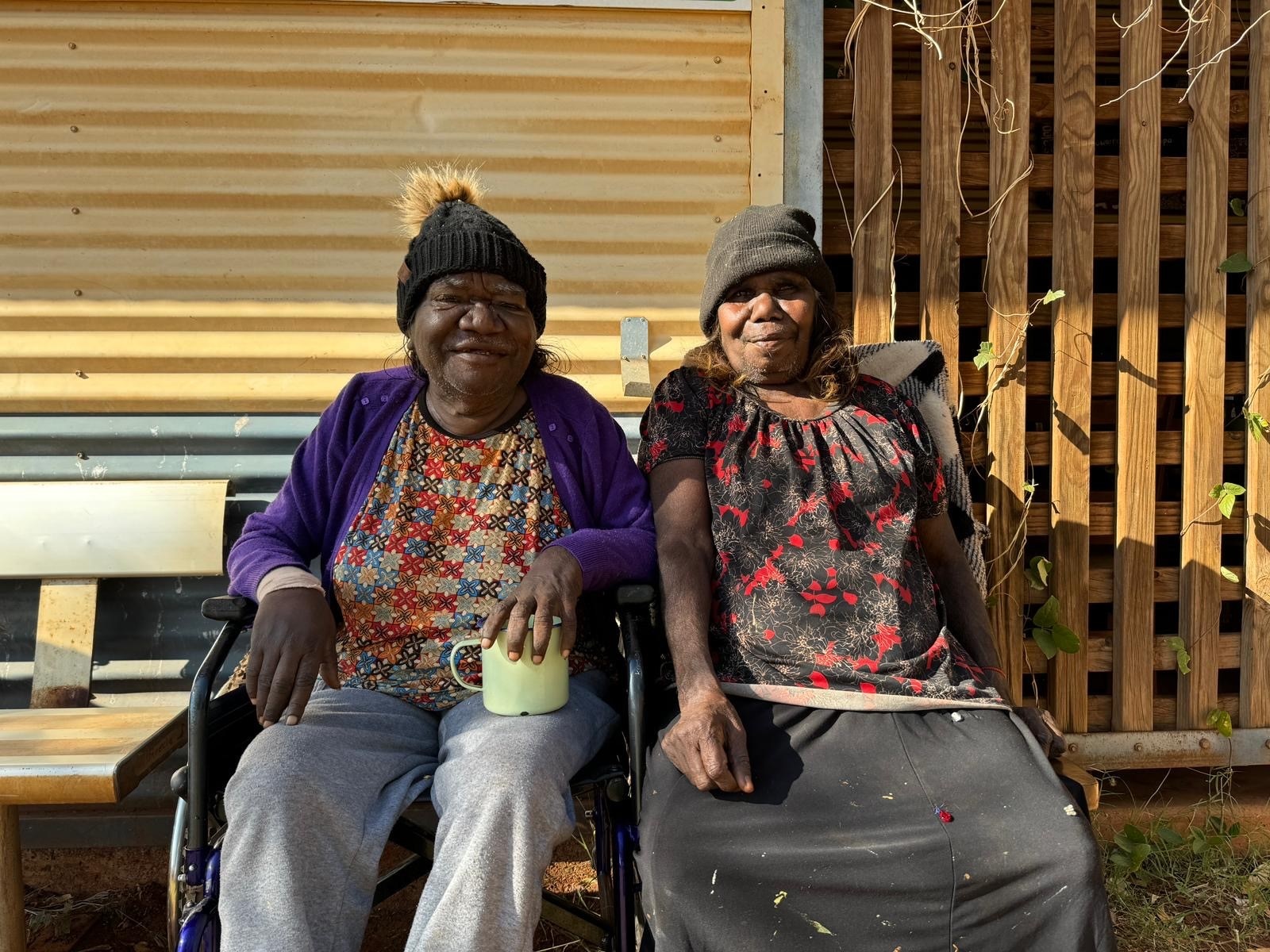 Two elderly women sitting on a bench and smiling, one woman is holding a cup of tea.