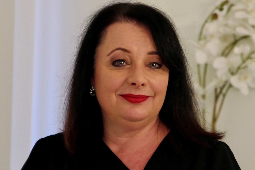 A head and shoulders shot of a woman with dark hair and red lipstick smiling for a photo indoors.