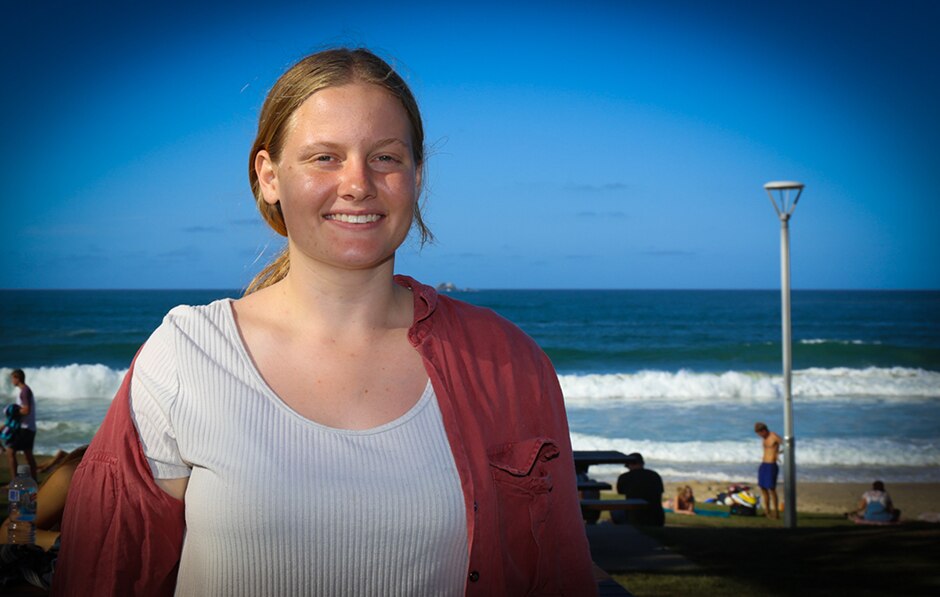 A young woman standing, with a beach and other beach-goers in the background.