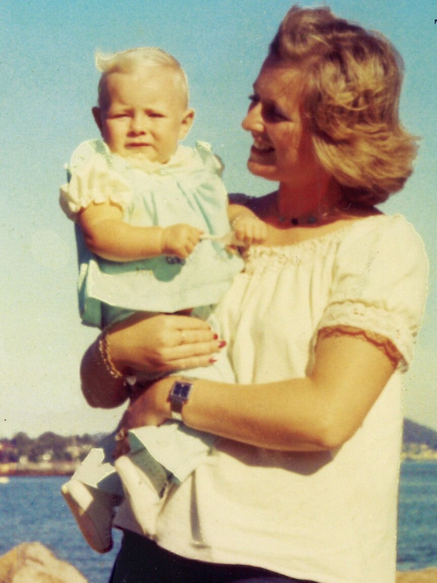 Young woman in1970s faashion adoringly gazes at a baby she holds in her arms at a beach