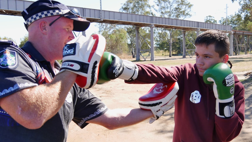 A teenage boy wearing green boxing gloves trains with a police officer in a sunny park in Bundaberg, Queensland.