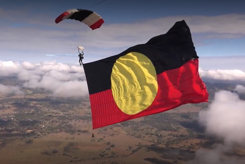 An Indigenous man skydives with a large Aboriginal flag flying behind him.