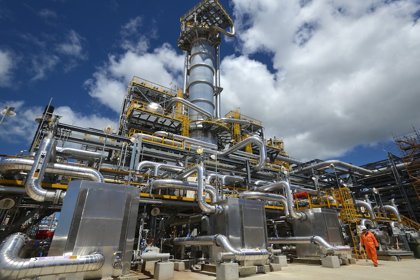 A person in orange overalls is dwarfed by a huge metal gas plant.