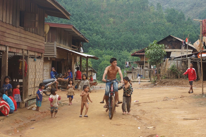 A man rides a bicycle while children play nearby on a muddy road