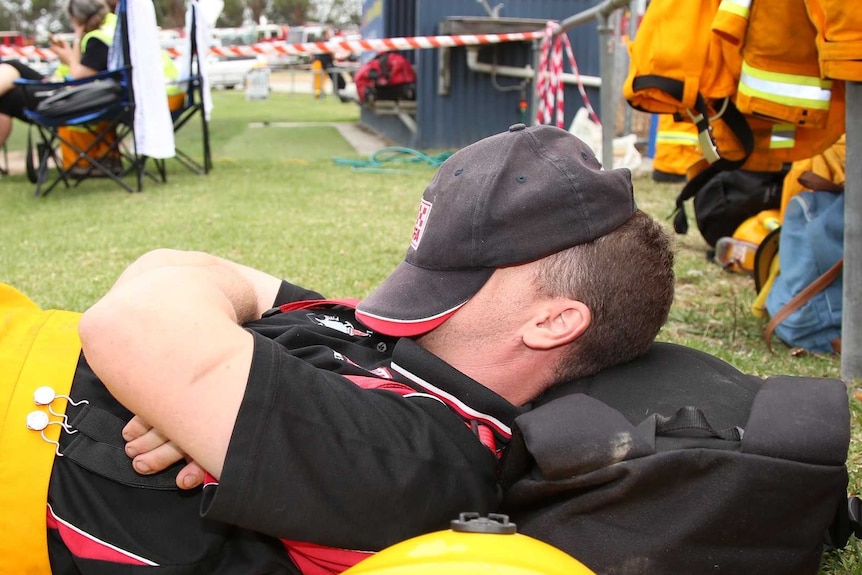 A man in yellow overalls lies against a backpack with a cap over his head.