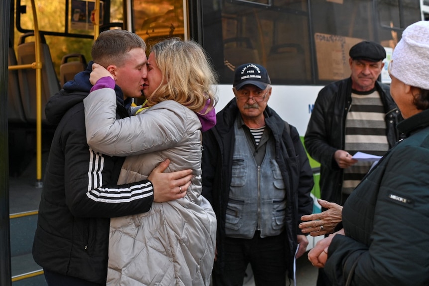 A man and a woman kiss and hug near a bus, as several people look on