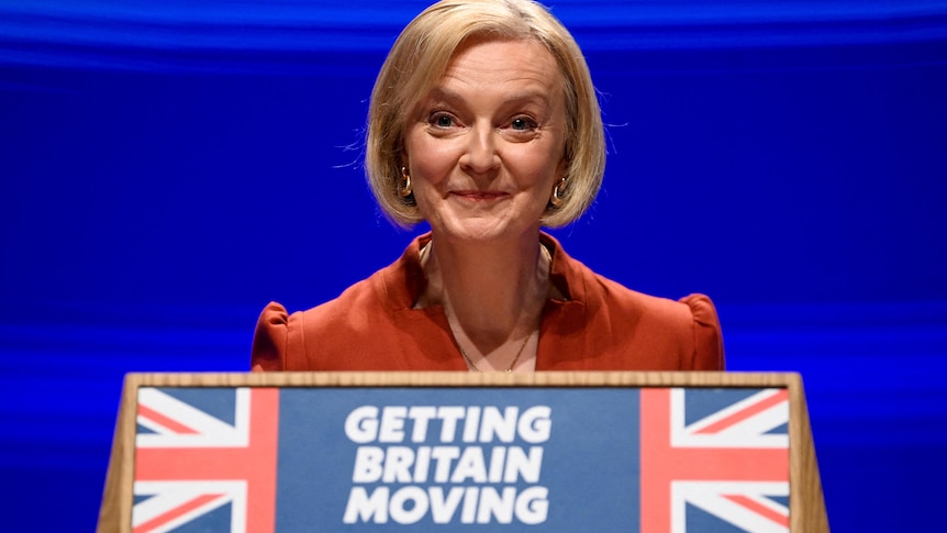 Liz Truss stands on stage and speaks behind a podium which reads 'Getting Britain Moving' with the Union Jack. 