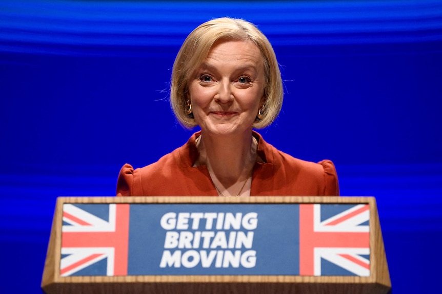 Liz Truss stands on stage and speaks behind a podium which reads 'Getting Britain Moving' with the Union Jack. 
