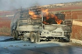 A burnt-out flat-bed truck sitting at the side of the road as fire continues to burn in its tray.