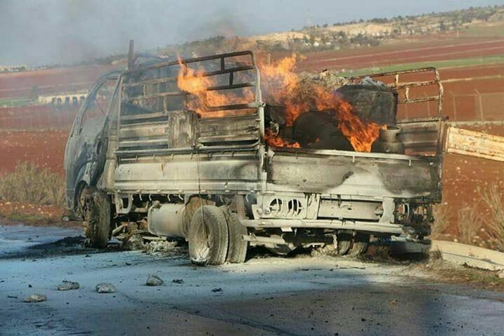 A burnt-out flat-bed truck sitting at the side of the road as fire continues to burn in its tray.