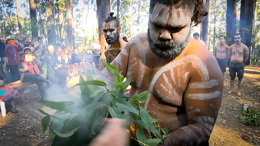 Two men in traditional Aboriginal face and body paint make their way around a circle of onlookers with burning leaves.