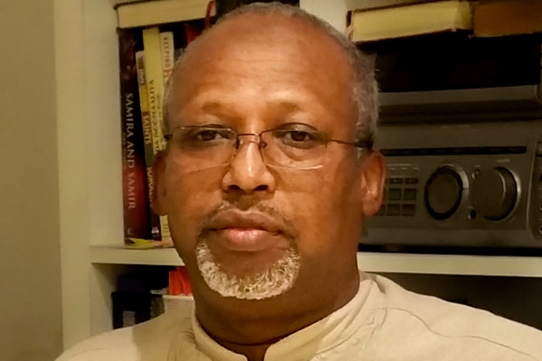 Dr Hussein Haraco, wearing a cream shirt and glasses, sits in front of a bookshelf.