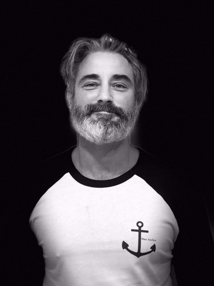 Black and white head and shoulders image of a man with grey hair wearing a t-shirt that says 'Man Anchor'.