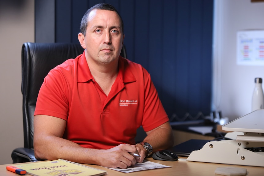 A man with short brown hair and in a red shirt sits at a desk with his hands crossed.