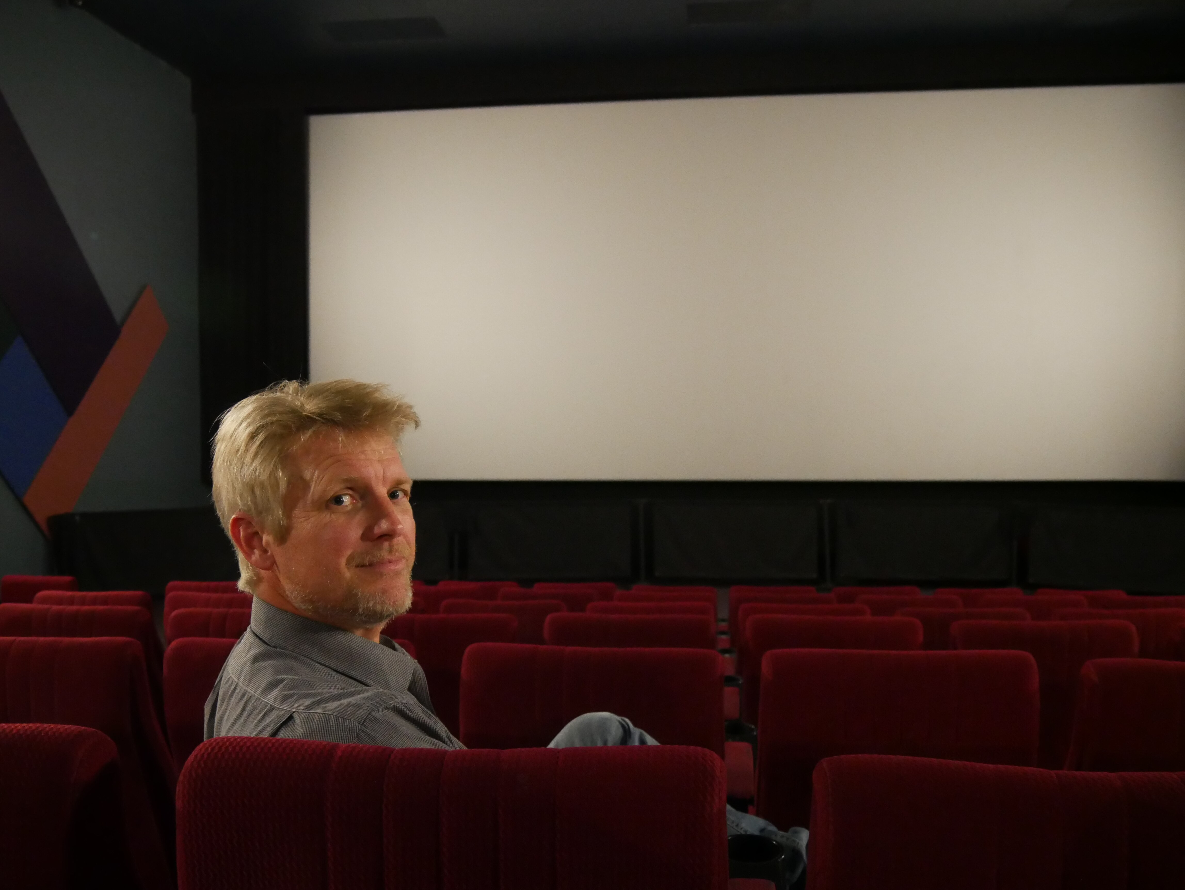 A man looking over the back of a cinema seat with a cinema screen behind him