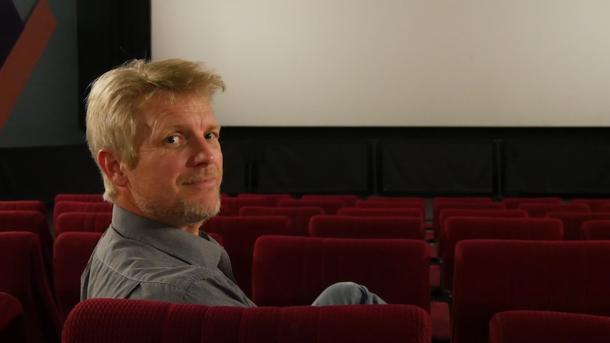 A man looking over the back of a cinema seat with a cinema screen behind him