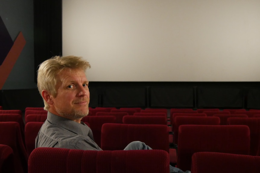 A man looking over the back of a cinema seat with a cinema screen behind him