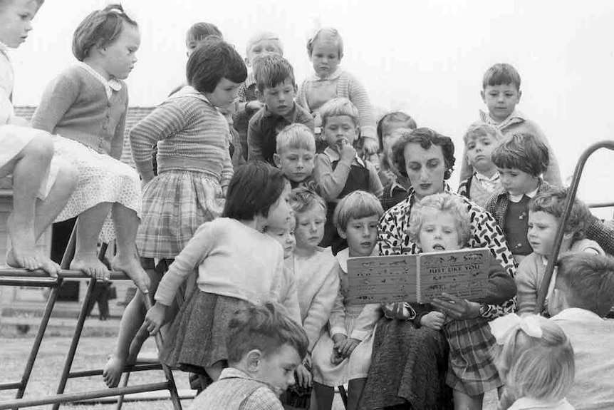 Children on play equipment listen to a woman reading a story.