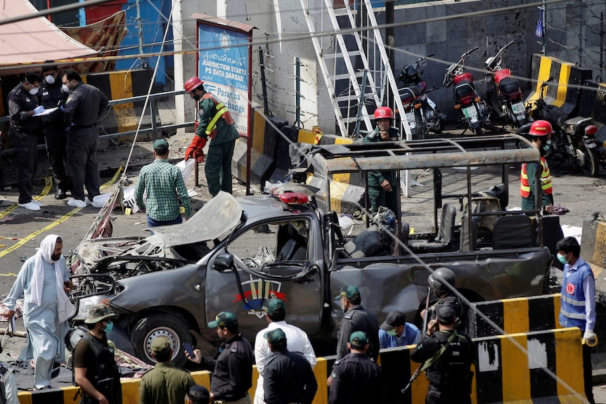 A truck is visibly damaged from a bomb blast. Some police crowd around something on the ground, others speak to each other.