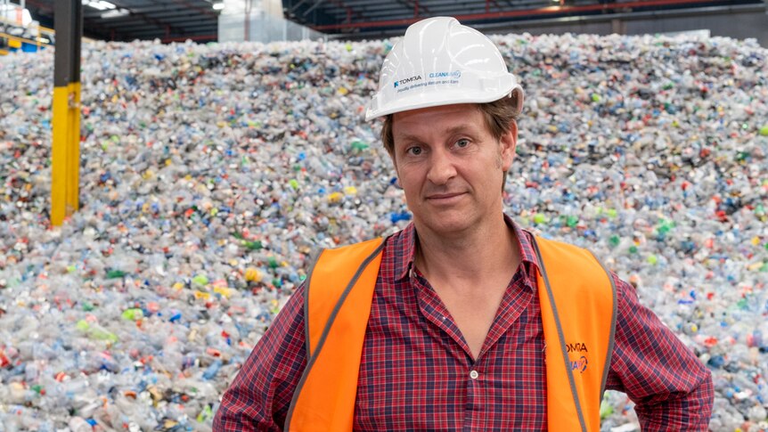 Craig Reucassel in a high vis orange vest and hard hat and standing in front of a pile of plastic bottles at a recycling plant