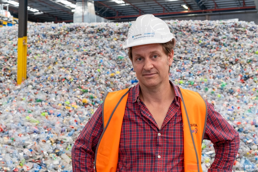 Craig Reucassel in a high vis orange vest and hard hat and standing in front of a pile of plastic bottles at a recycling plant