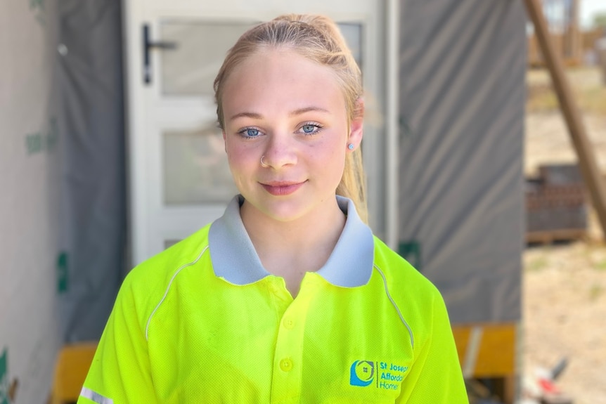 A girl smiles while wearing a fluorescent tradie top and standing in front of a construction site.