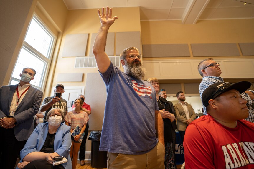 A man with a long beard in an American flag t-shirt raises his hand in a crowd 
