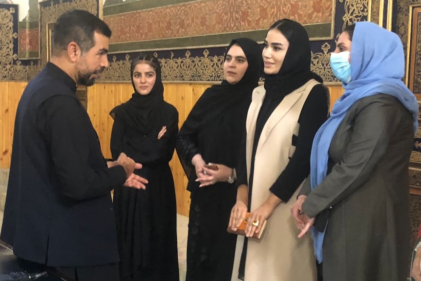 A man speaks to three women wearing head scarves and conservative dress in an official setting.