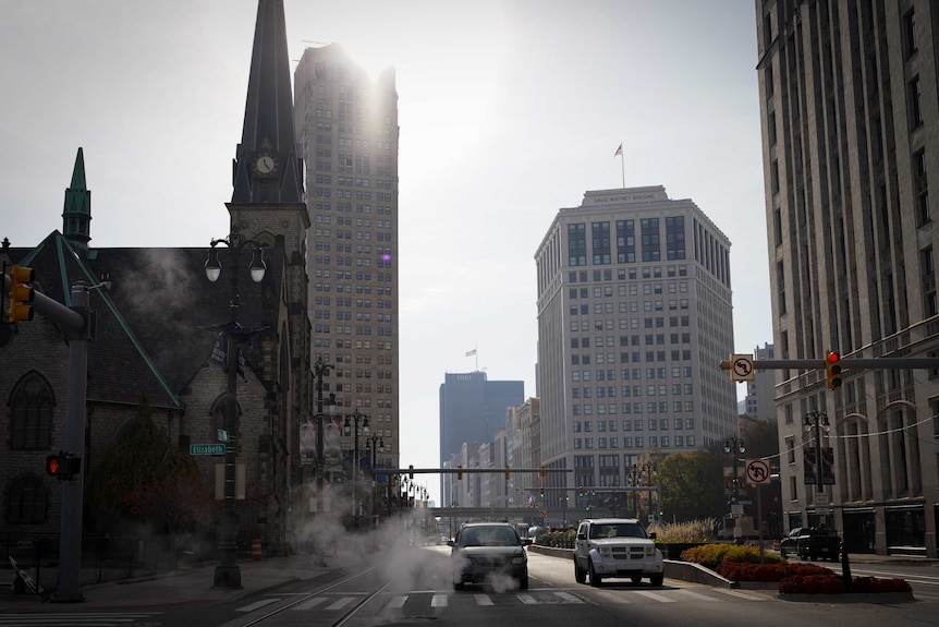 Two cars wait at traffic lights on a street as smoke blows by against a city backdrop.