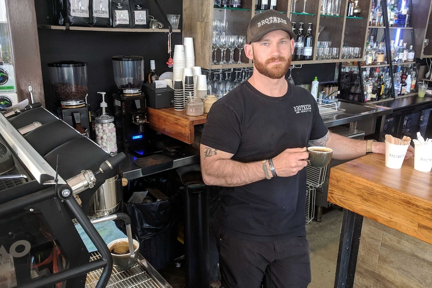 A man wearing a black t-shirt and cap with the logo 'Brothers' stands behind a counter holding a cup of coffee