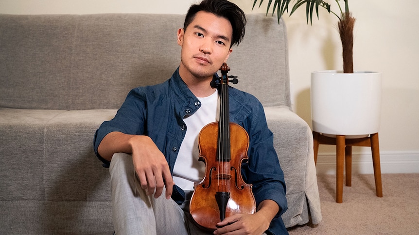 Ray Chen sits on the floor in front of a grey lounge holding a violin resting on his leg upright