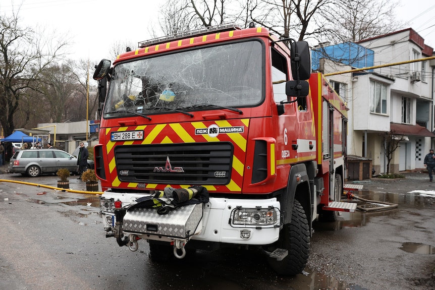 A fire truck with a smashed window is parked in a street with damaged houses in the background