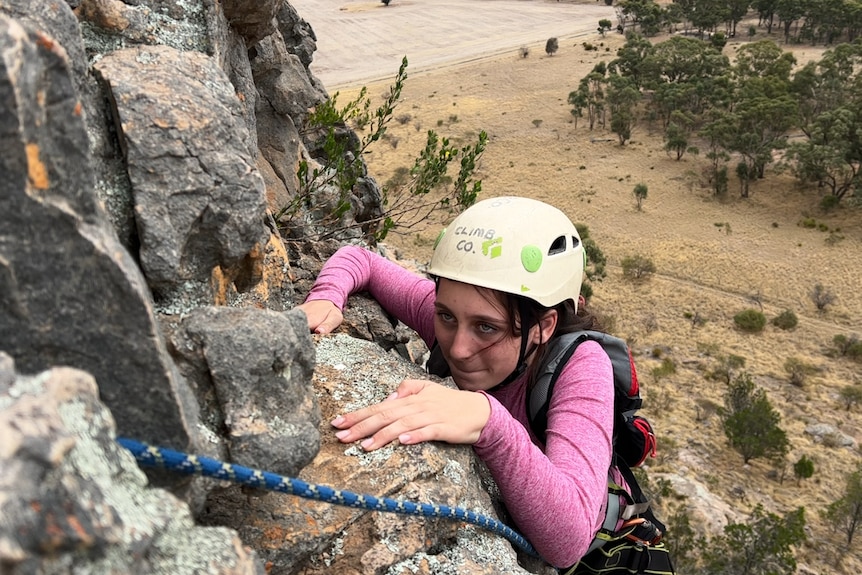 A girl wearing a pink shirt and white helmet pushes herself up a rock face with a determined look. a blue rope in the foreground