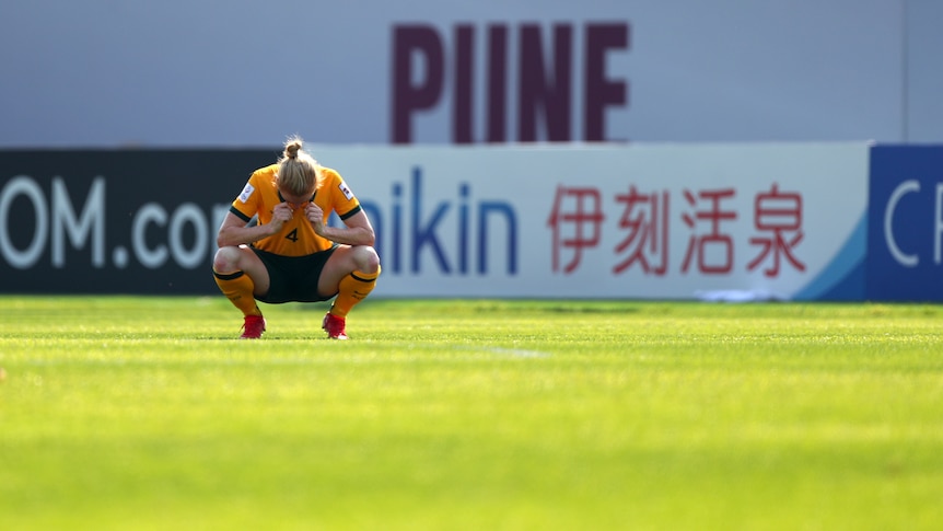 Australian soccer player leans down on her haunches and wipes away tears