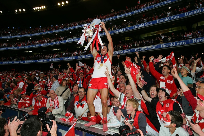 A helmet-wearing AFL player stands on a fence at the MCG hoisting the premiership cup as the crowd cheers behind him