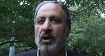 A screen grab of a man wearing a grey suit talking to tv cameras.