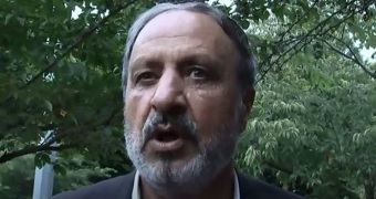A screen grab of a man wearing a grey suit talking to tv cameras. A screen grab of a man wearing a grey suit talking to tv cameras.