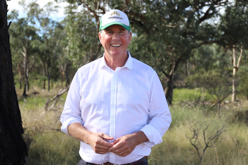 Man in white shirt with hat on smiles whilst standing in front of bushland