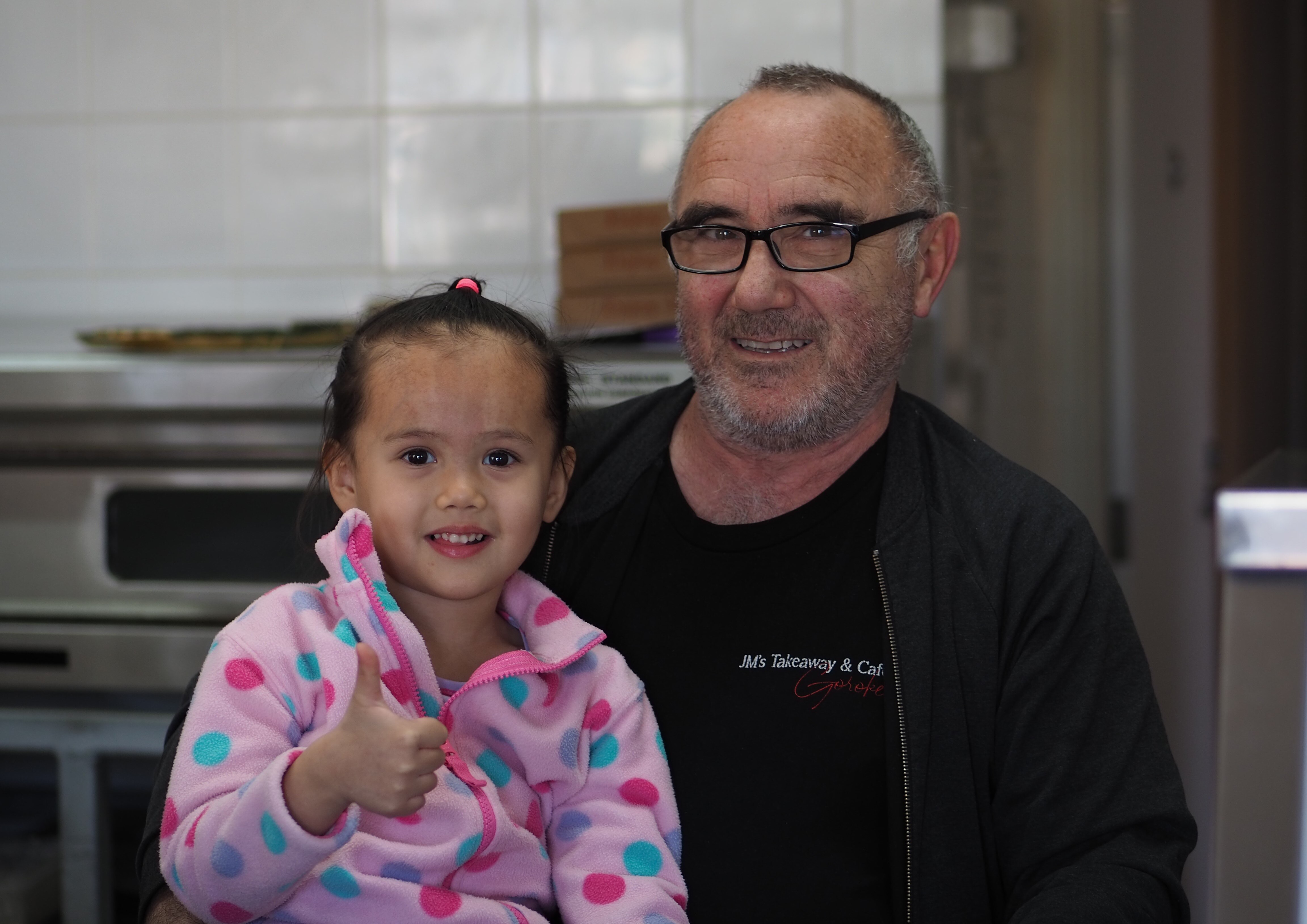 Una niña con un mono rosa levanta el pulgar. Su padre, al fondo, sonríe a la cámara. Al fondo, una tienda.