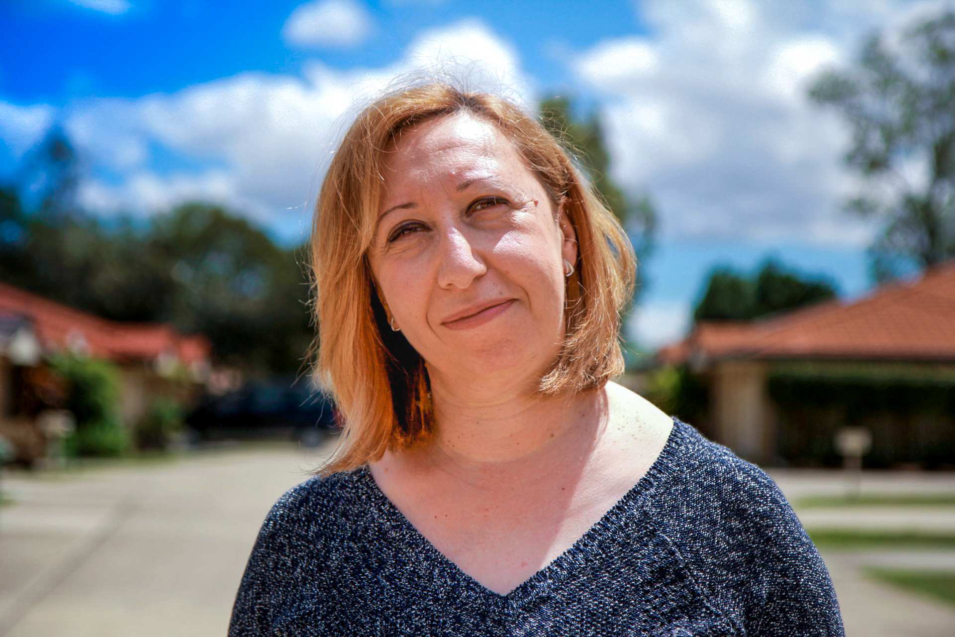 A profile shot of a woman with short blonde-brown hair standing in front of an out-of-focus suburban background.